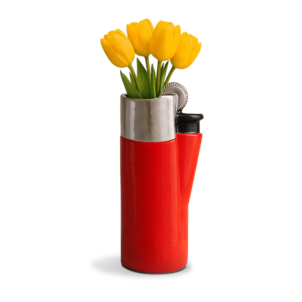 A novelty vase shaped like a red lighter with a silver metallic top and a black nozzle, holding yellow tulips against a white background.